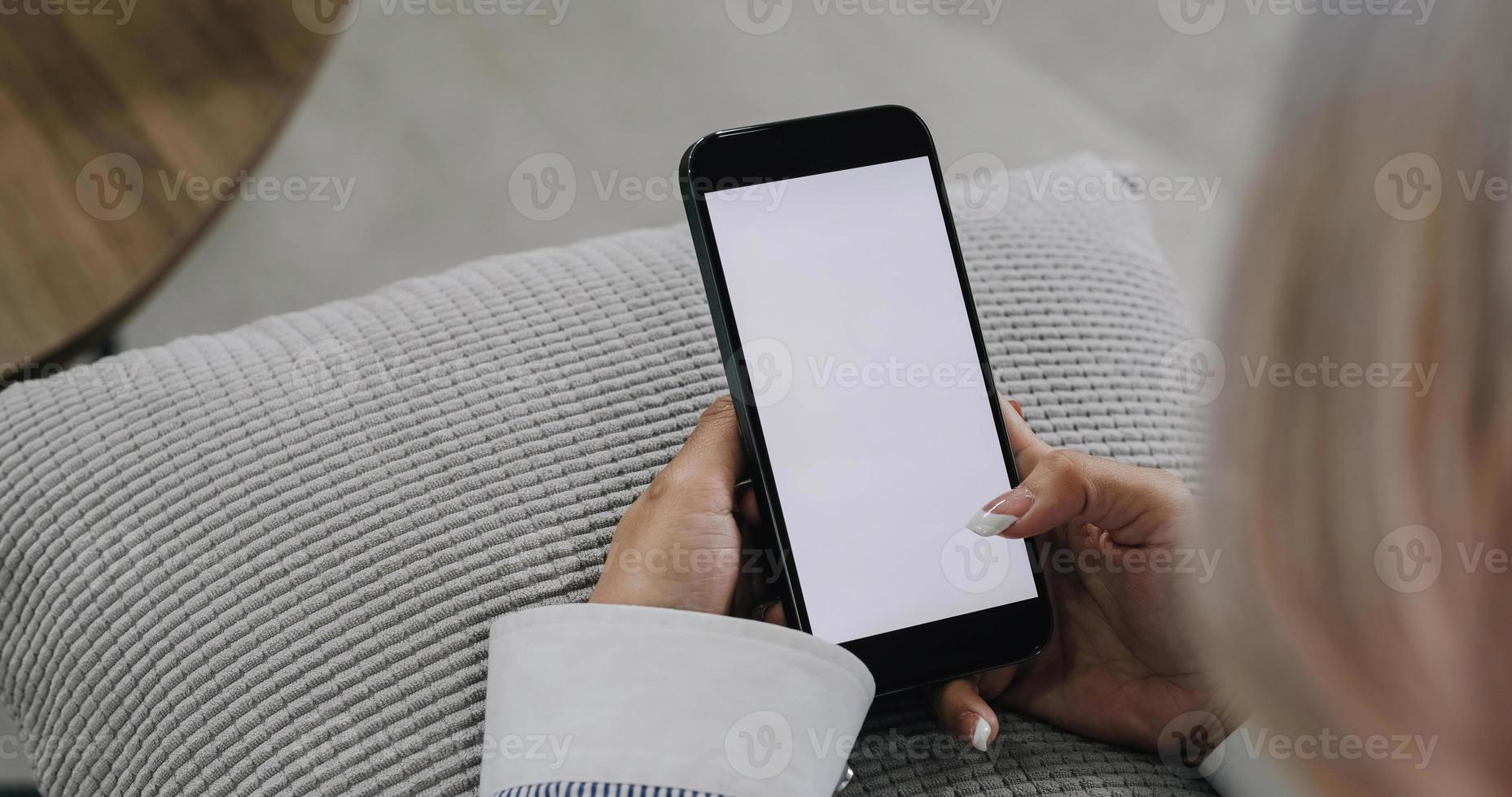 imagen de primer plano, hermosa joven asiática usando su smartphone mientras se relaja en su sala de estar mínima. una mujer sosteniendo una maqueta de pantalla blanca de teléfono móvil. foto