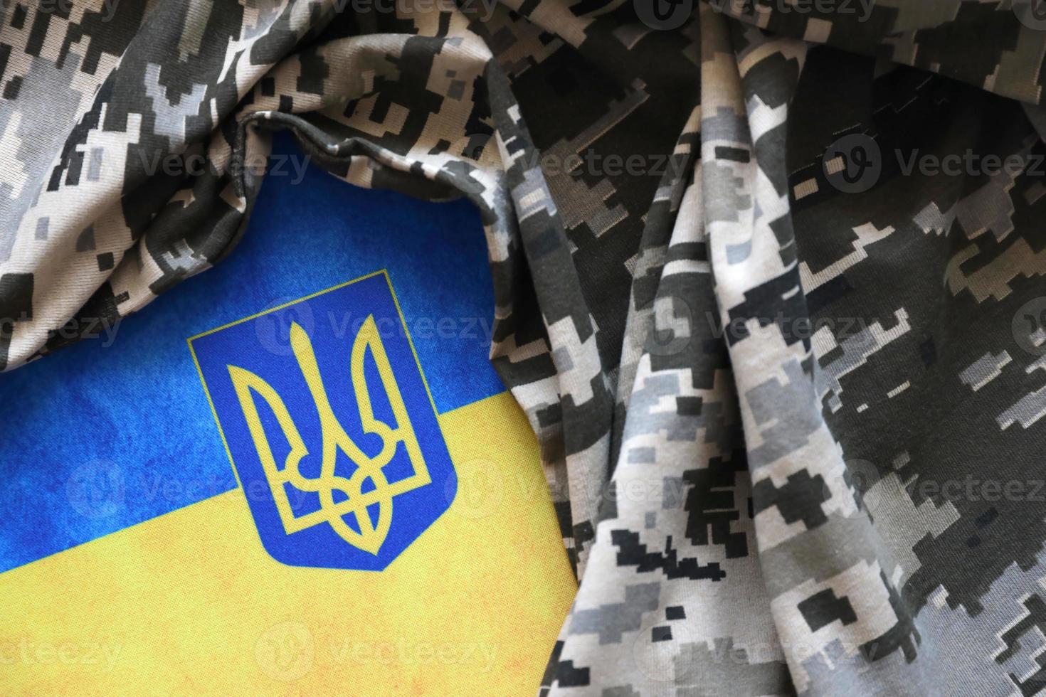 Ukrainian flag and coat of arms with fabric with texture of pixeled camouflage. Cloth with camo pattern in grey, brown and green pixel shapes with Ukrainian trident sign photo