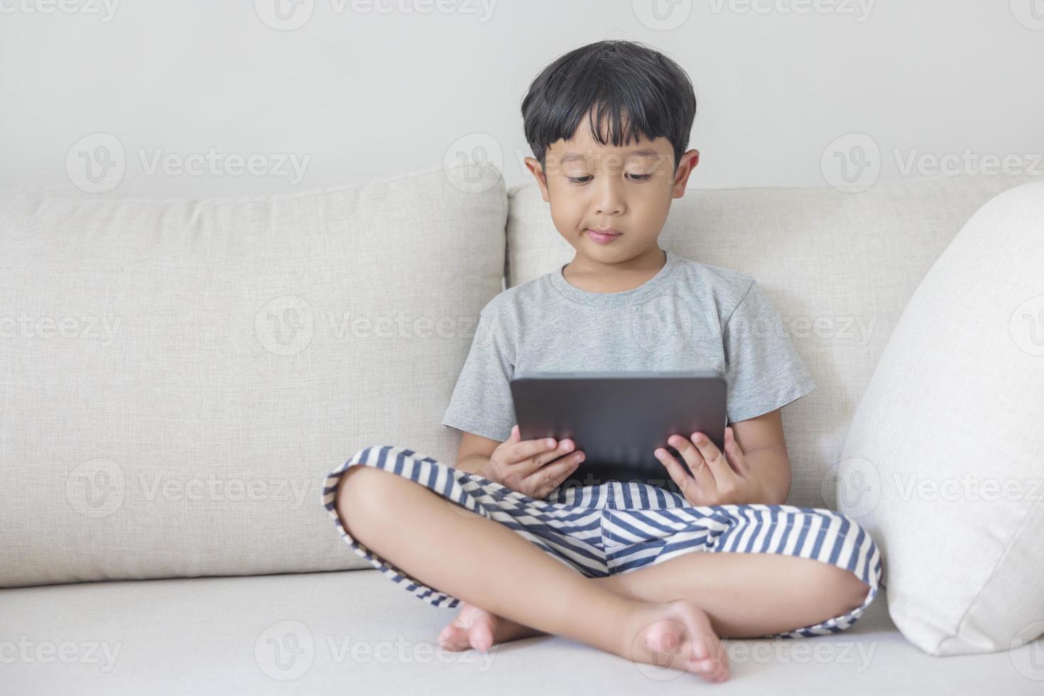 Adorable happy Asian boy wearing a gray shirt and blue-white striped shorts is having fun playing with his tablet on a cream sofa. looking at the mobile screen photo