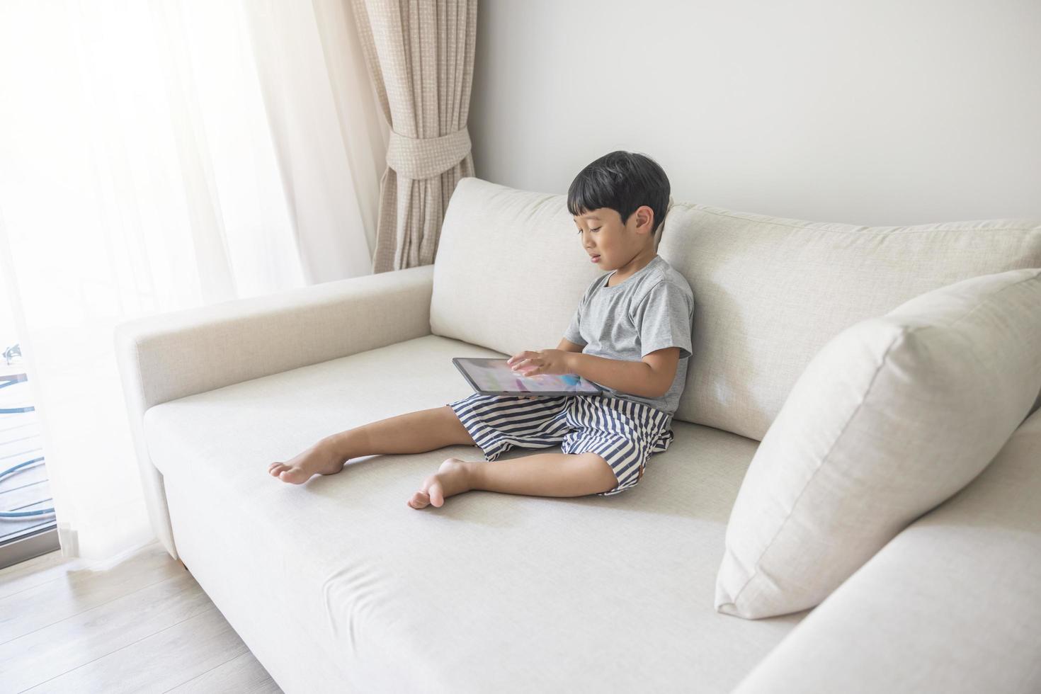 Adorable happy Asian boy wearing a gray shirt and blue-white striped shorts is having fun playing with his tablet on a cream sofa. looking at the mobile screen photo