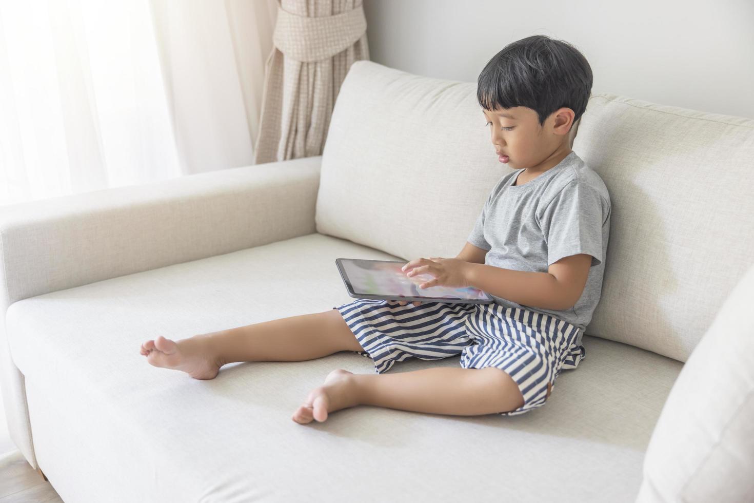 Adorable happy Asian boy wearing a gray shirt and blue-white striped shorts is having fun playing with his tablet on a cream sofa. looking at the mobile screen photo
