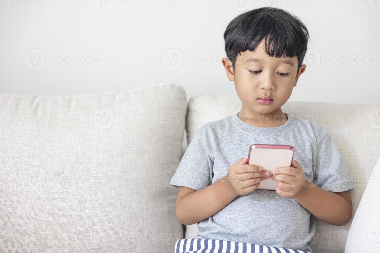 An adorable happy Asian boy wearing a gray shirt and blue-white striped shorts is having fun playing with his smartphone on a cream sofa. looking at the mobile screen photo