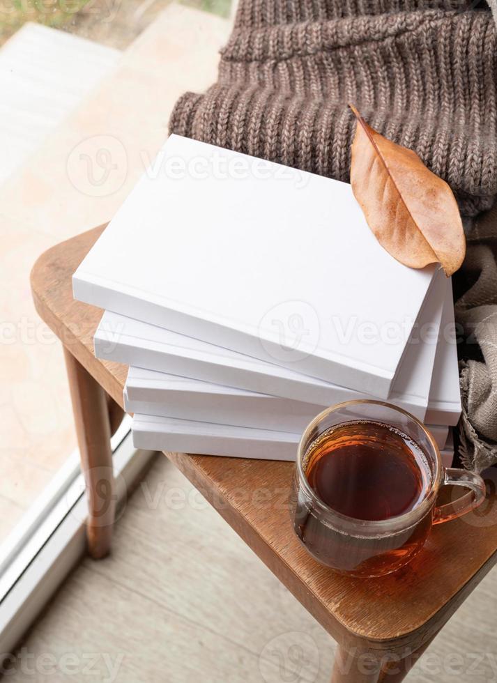 Stack of white blank books with autumn leaves and cup of hot tea on old wooden chair, mockup design photo