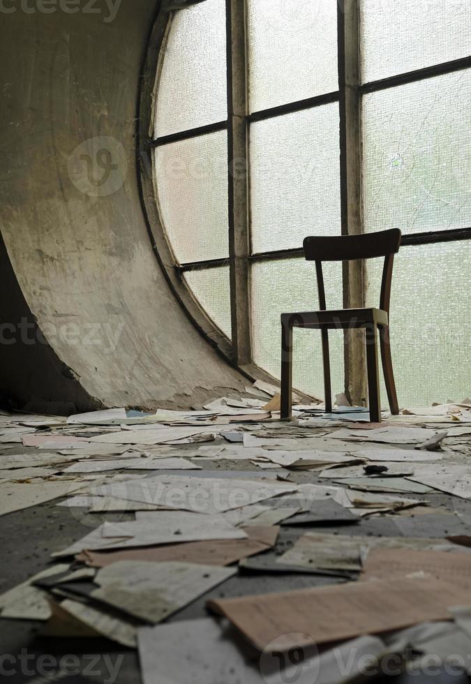 Chair in front of a round window in an abandoned church with papers and book pages lying on the ground photo