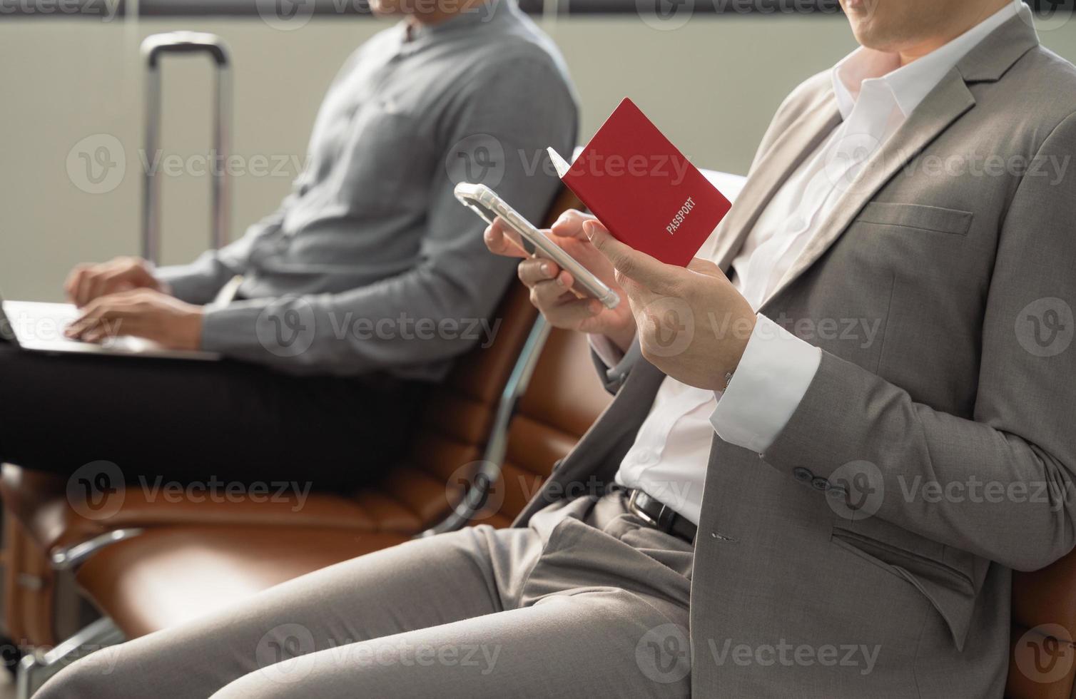 Businessman holding passport sitting at airport terminal and checking for his flight on mobile phone, waiting for plane and working while travelling photo