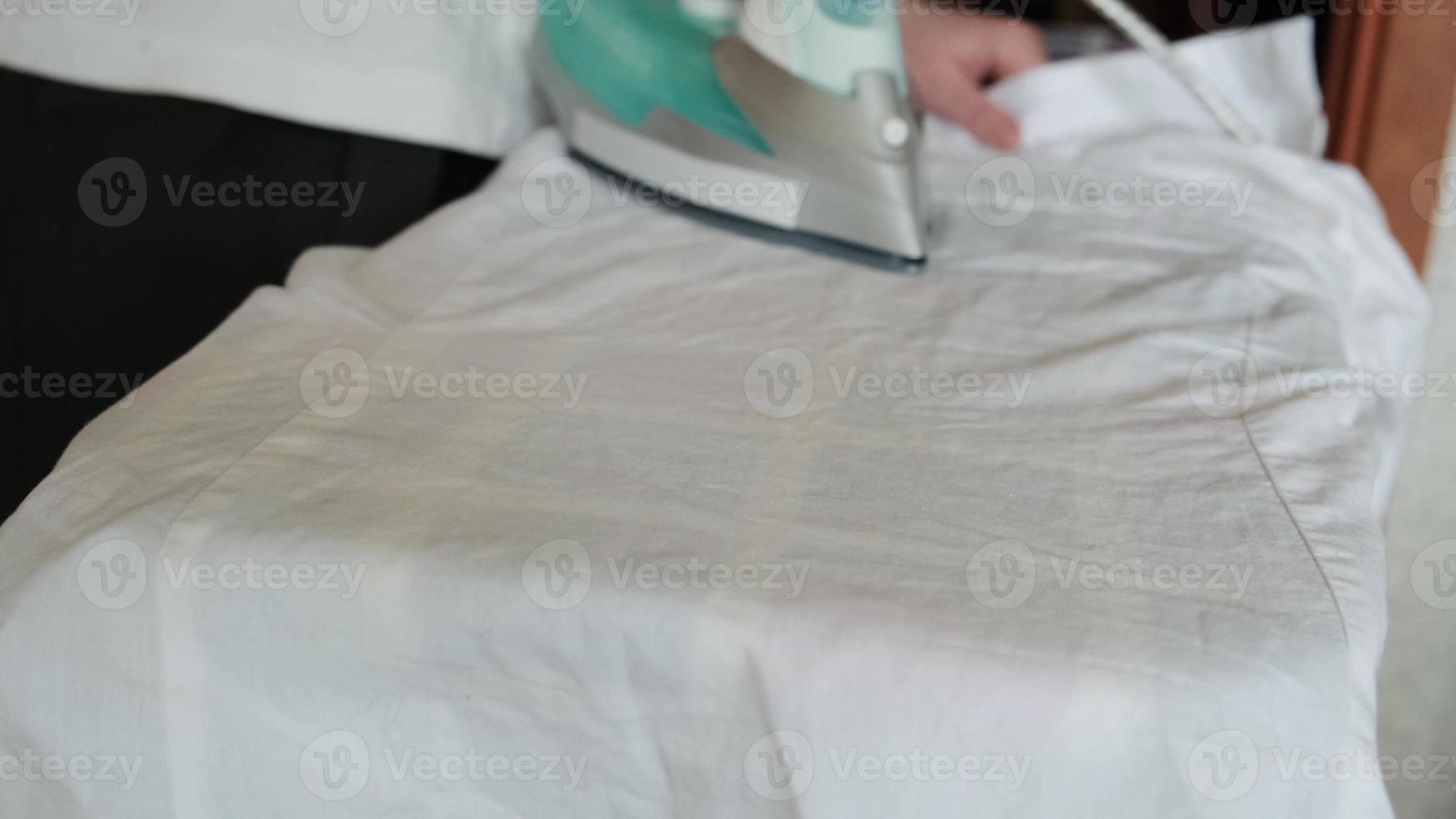 Female hand holding electric iron ironing clothes on ironing board. Woman ironing white clothes closeup. Home routine housekeeping housework household chores. Maid working with clean clothing. photo