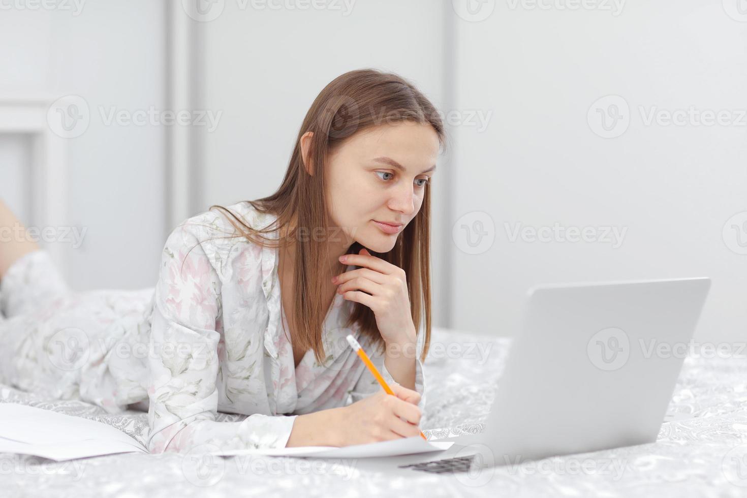 Beautiful young woman working, using laptop computer and makes notes in the bedroom at home. Freelancer. Writing, typing. Girl checking social apps. Communication and technology concept photo