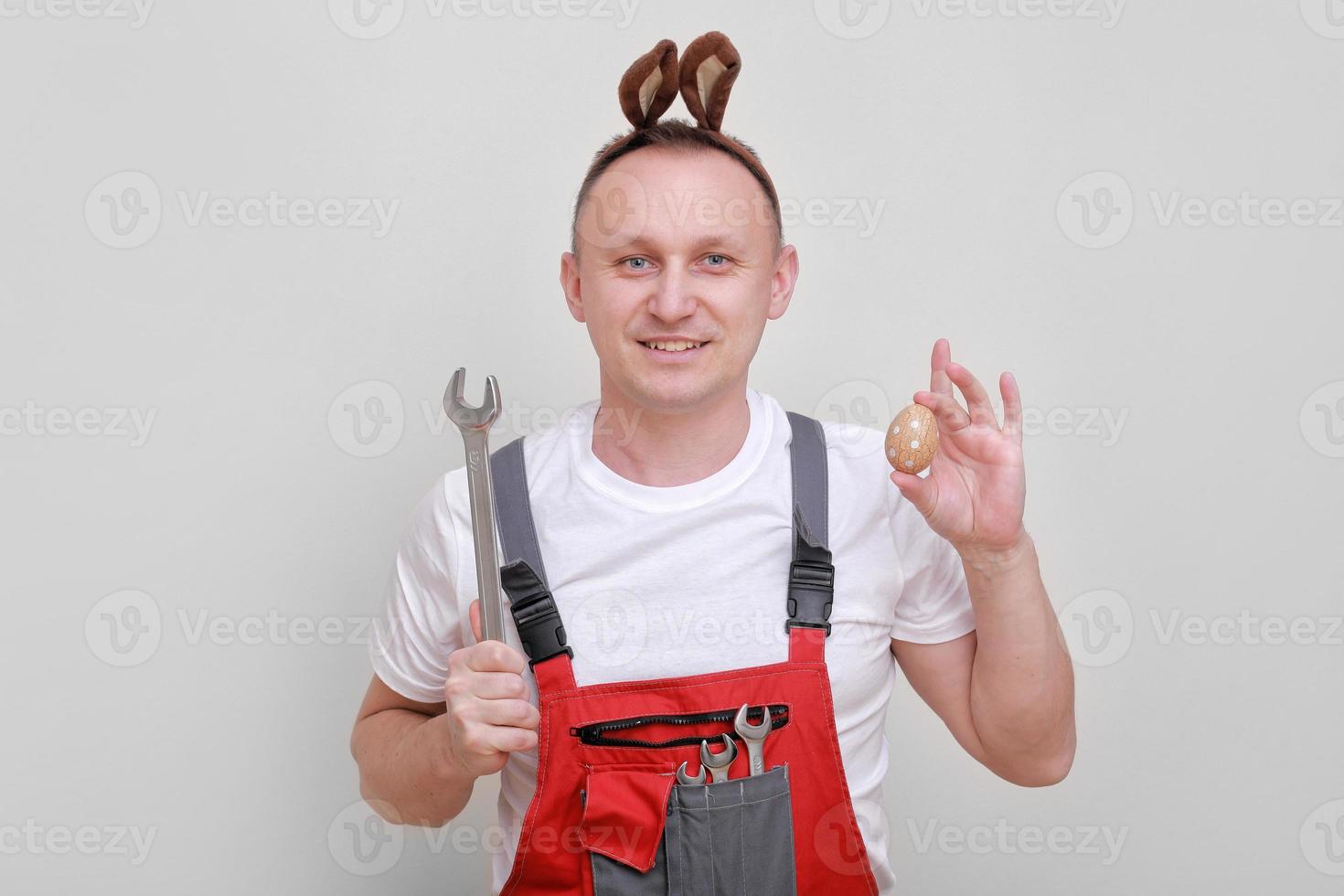 Easter holiday, celebration concept. funny smiling engineer worker or mechanic is wearing rabbit ears on head, holding wrenches and painted egg white background. celebrating orthodox day photo