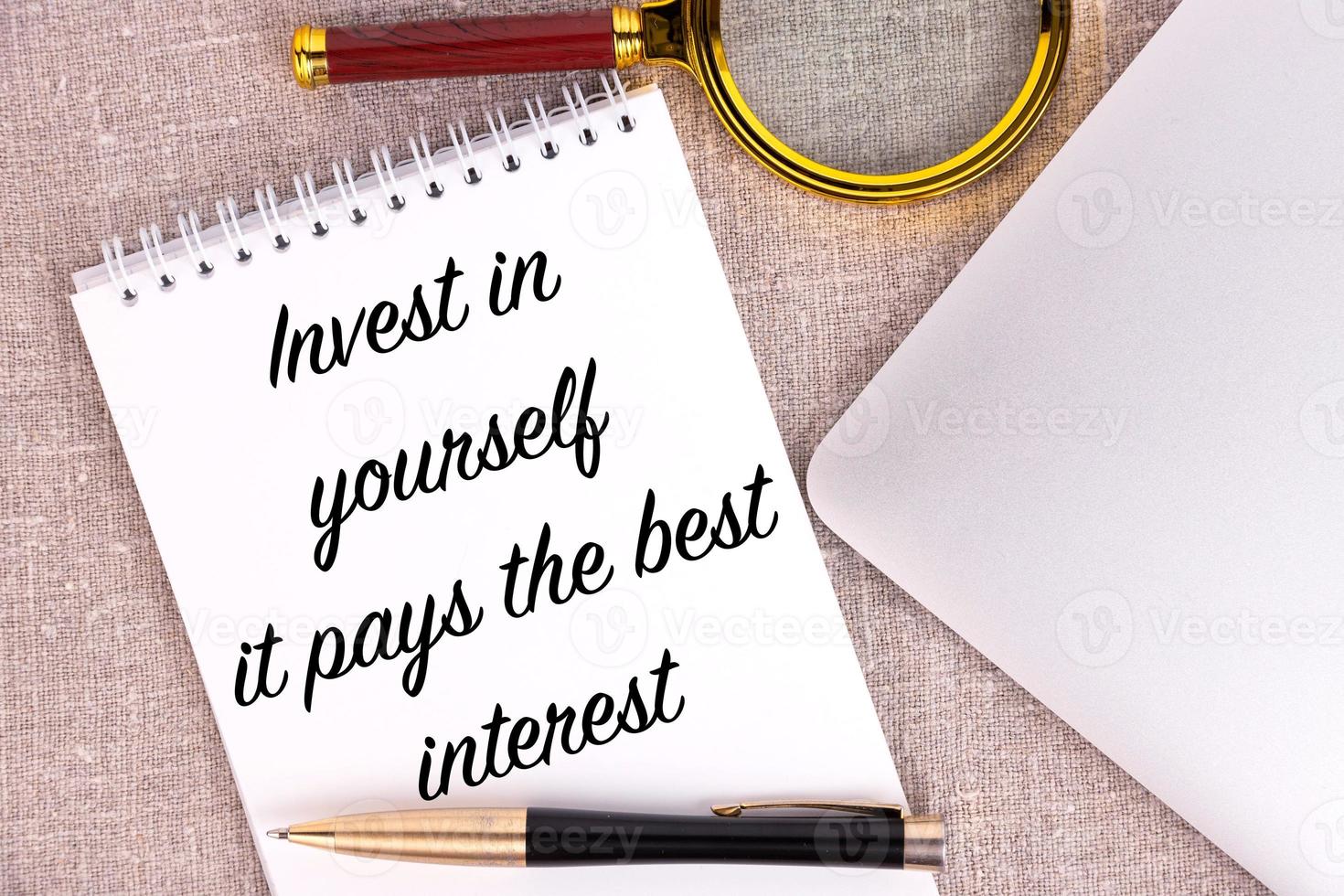 Invest in yourself it pays the best interest, the text is written in a notebook, next to a pen, a magnifying glass and a laptop on a linen background. photo