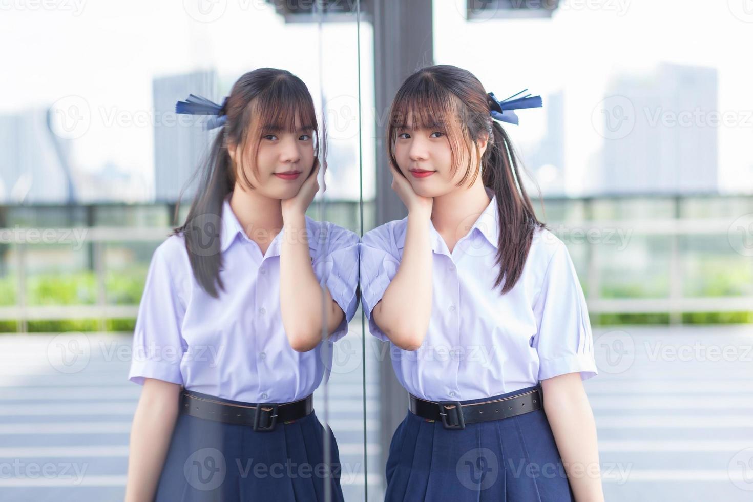 Beautiful Asian high school student girl in the school uniform with stands smiles confidently beside the mirror while she looks at the camera happily with city in the background. photo
