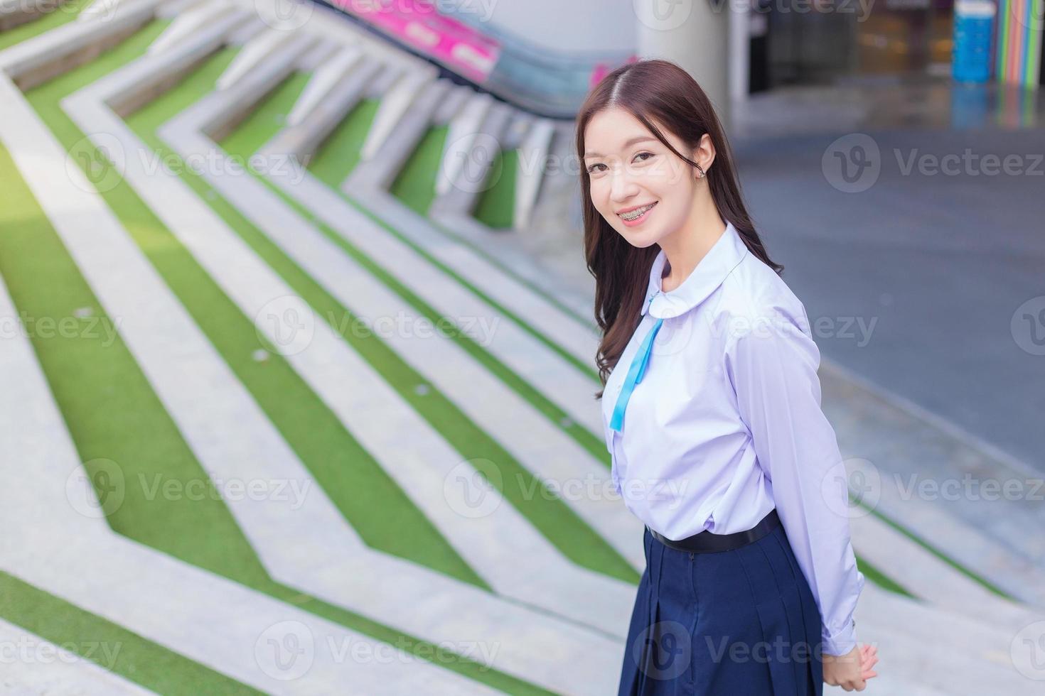 Beautiful high school Asian student girl in the school uniform stands and smiles happily with braces on her teeth while she put her hand on head confidently with the building as a background. photo