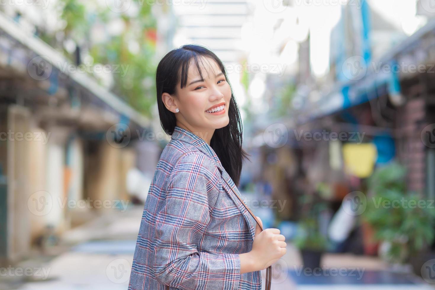 Confident young Asian woman who wears a Brown striped blazer and shoulder bag smiles happily and looks at the camera as she commute to work through the old town. photo