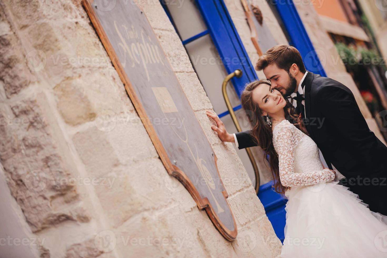 pareja de novios abrazándose en la ciudad vieja. puertas vintage azules y café en la antigua ciudad de fondo. novia elegante con vestido largo blanco y novio con traje y corbata de moño. día de la boda foto