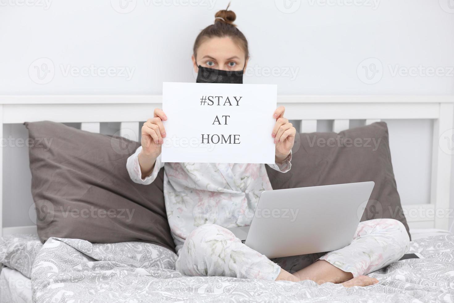 Coronavirus. Young woman in protection mask with laptop holding paper with Stay Home on bed. isolated to prevent infection of pandemic, epidemic Covid-19. Quarantine at home. selective focus. photo