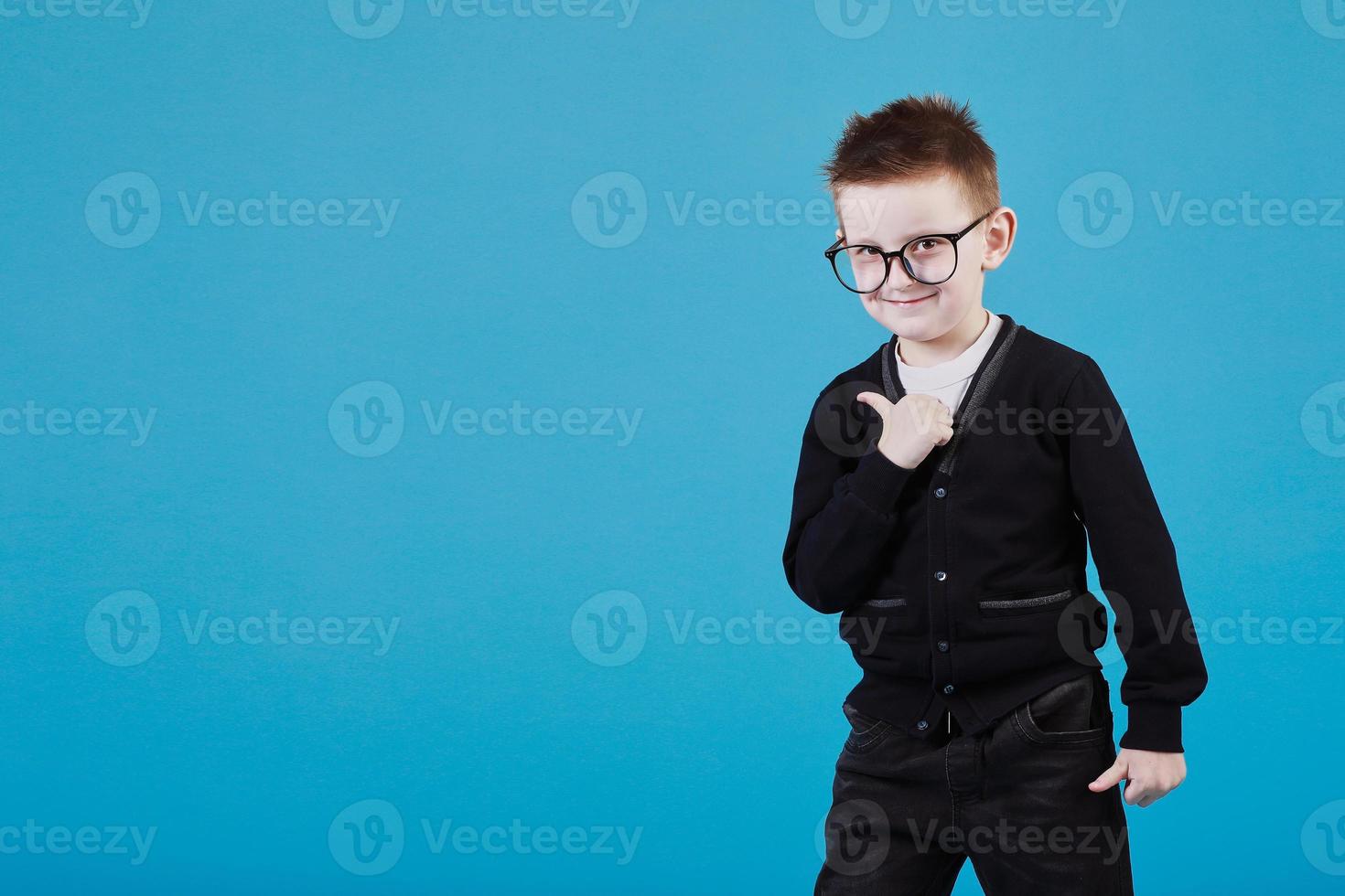 Like. Portrait of happy little schoolboy with glasses smiling at camera and doing thumbs up gesture, showing agree cool approval sign. indoor studio shot isolated on blue background photo