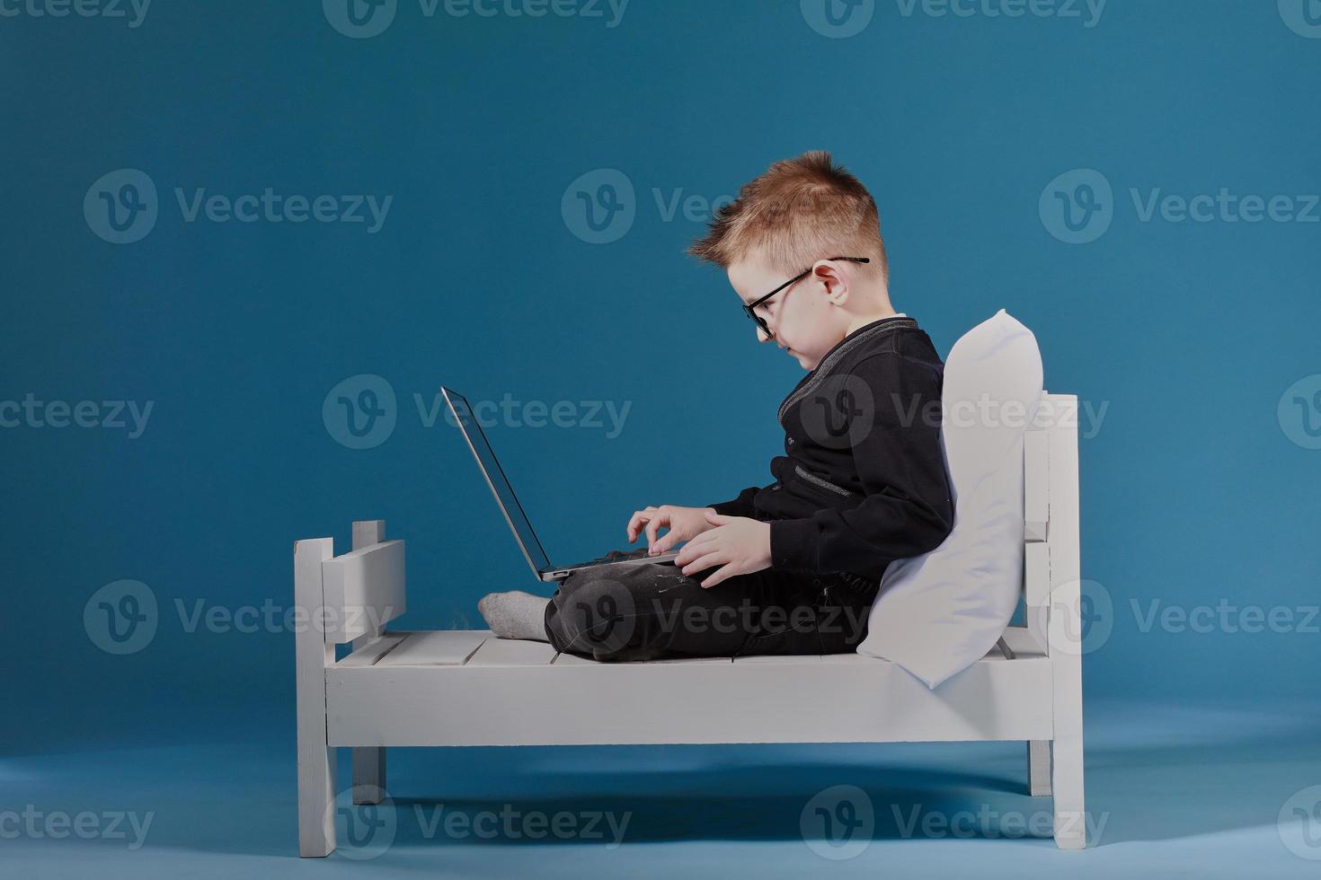 schoolboy in glasses using laptop sitting on bed. Child doing school homework. Online learning, distance lesson, education with online tutor at home isolation photo