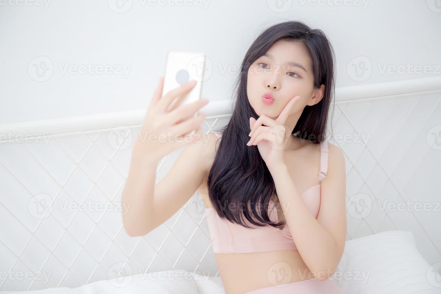 Beautiful young asian woman sexy in underwear talking a selfie on smartphone for social network in the bedroom, girl in lingerie relax with taking a picture on mobile phone on bed in the bedchamber. photo