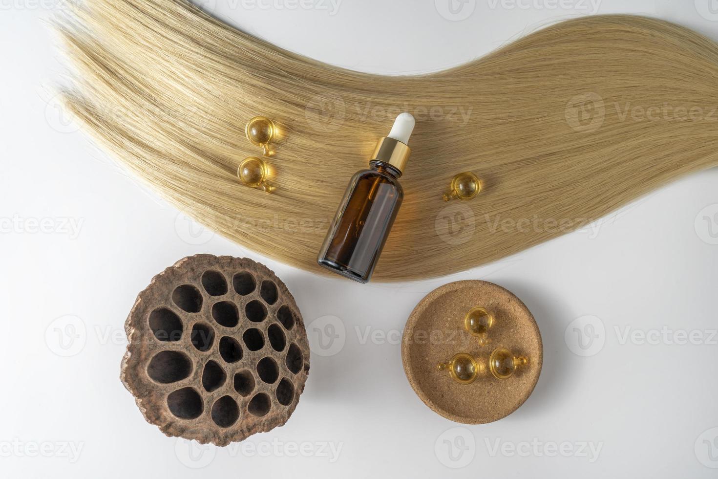 A hair care oil or serum in a brown dropper bottle and golden capsules lying on a blond hair strand, product marketing mockup photo