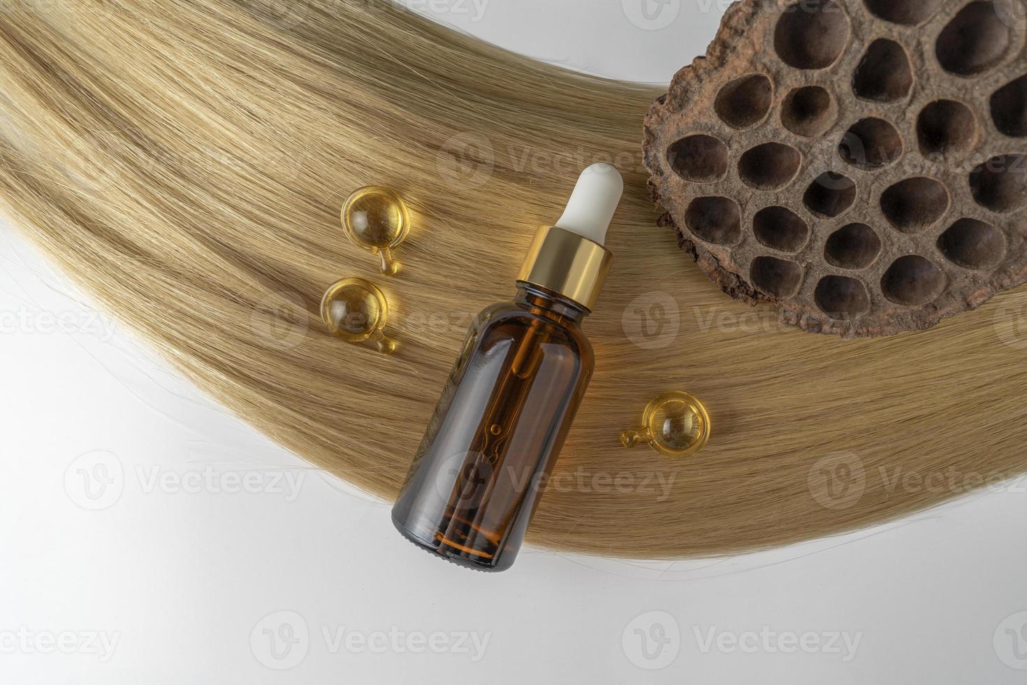 A hair care oil or serum in a brown dropper bottle and golden capsules lying on a blond hair strand, product marketing mockup photo