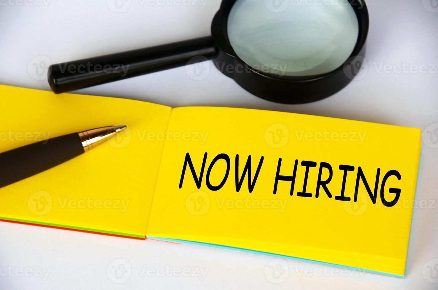 Now hiring text on yellow notepad with magnifying glass and pen on white background. Employment concept photo