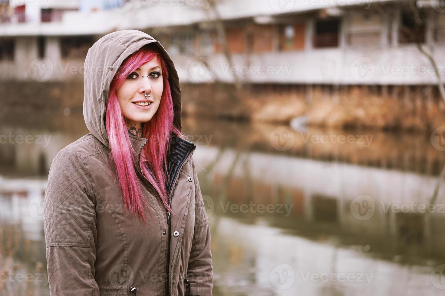 mujer joven perforada con cabello rosado y parka encapuchada de pie junto al río foto