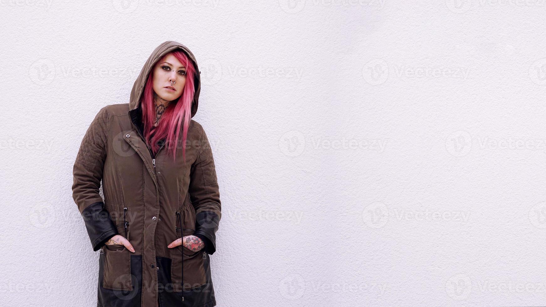young woman with hooded parka pink hair piercings and tattoos against wall with copy space photo