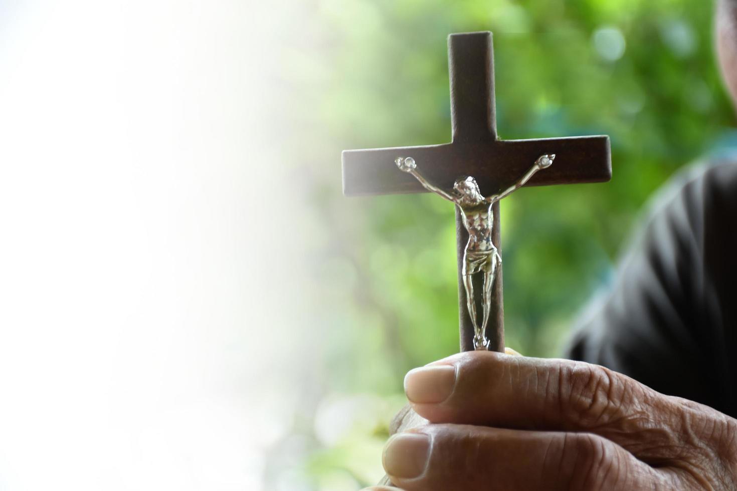 Closeup wooden cross which has a metal statue of crucified Jesus is in the hands of an asian eldery Catholic while praying in a local church, soft and selective focus. photo