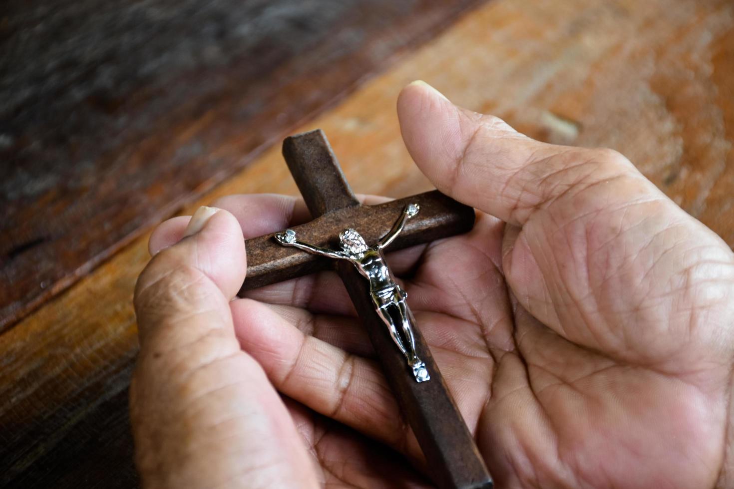 Closeup wooden cross which has a metal statue of crucified Jesus is in the hands of an asian eldery Catholic while praying in a local church, soft and selective focus. photo