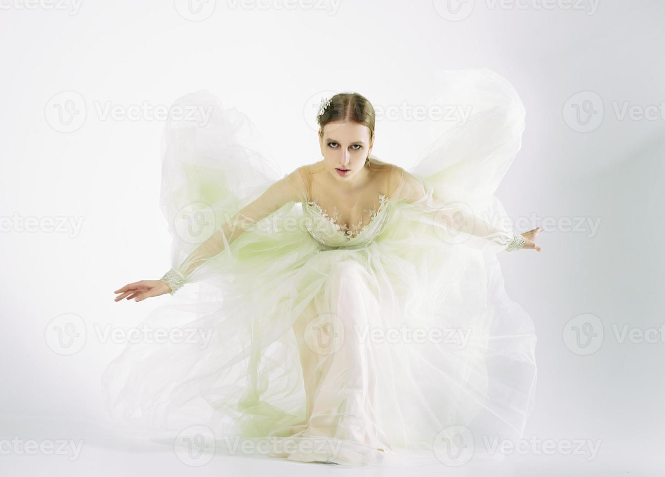 young girl model in a photo studio in a wedding dress sitting on one knee playing with a dress