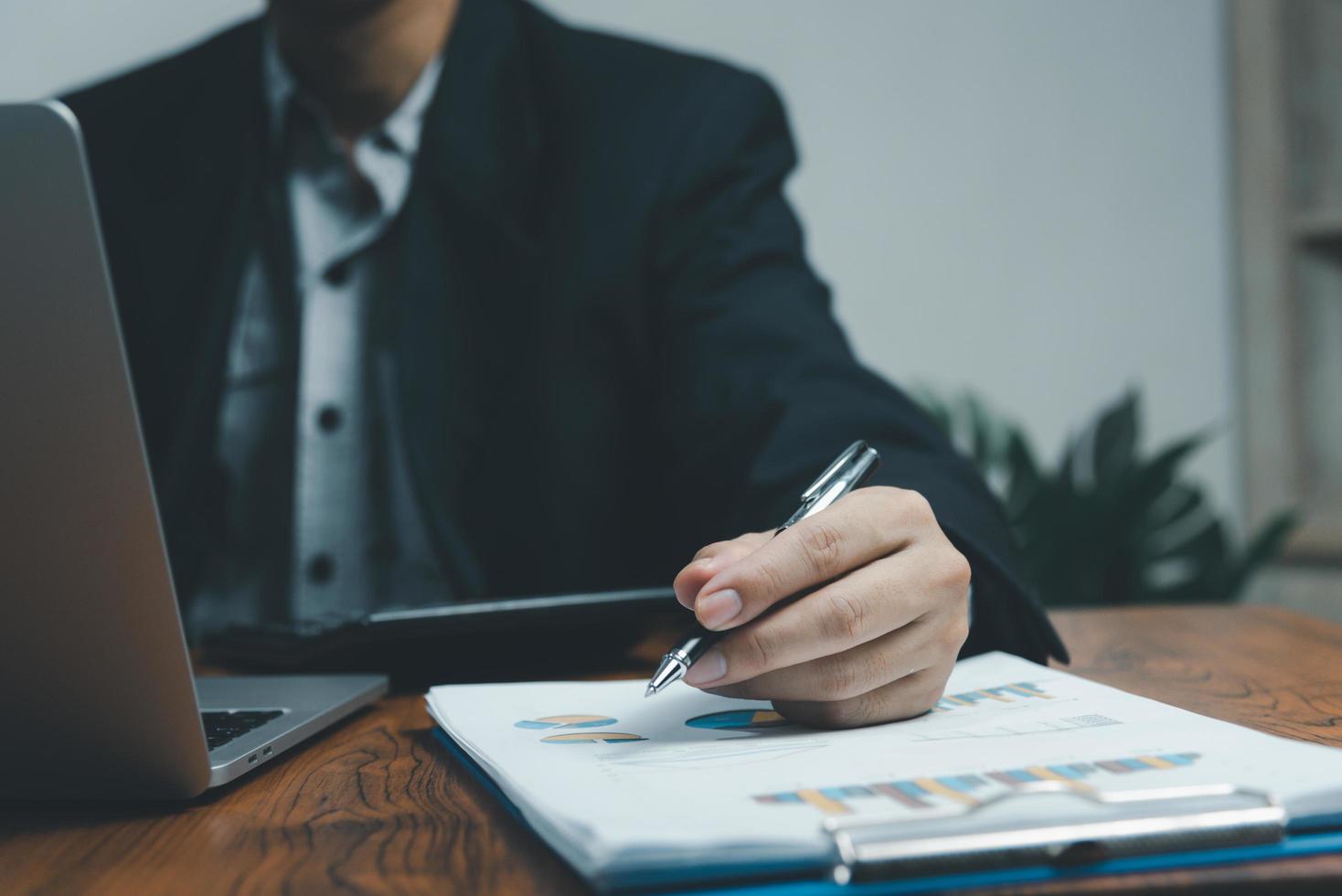 Businessman holding pen document graph accounting finance paper work analysis chart marketing investment and report economy with computer laptop on desk at office. photo
