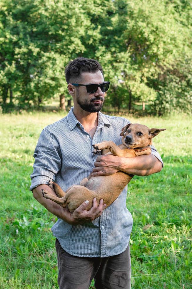 Handsome stylish European man is holding his dog in hands in park on a walk. Friendship between human and pets. photo