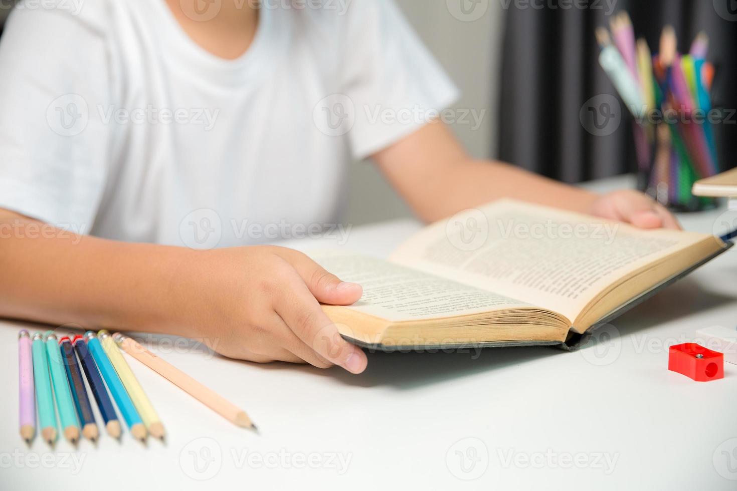 niño sentado estudiando en casa, niño estudiante en línea aprendiendo y haciendo deberes en el escritorio, niño pequeño leyendo y escribiendo un libro en la mesa. concepto de educación, ciberespacio tecnológico. foto