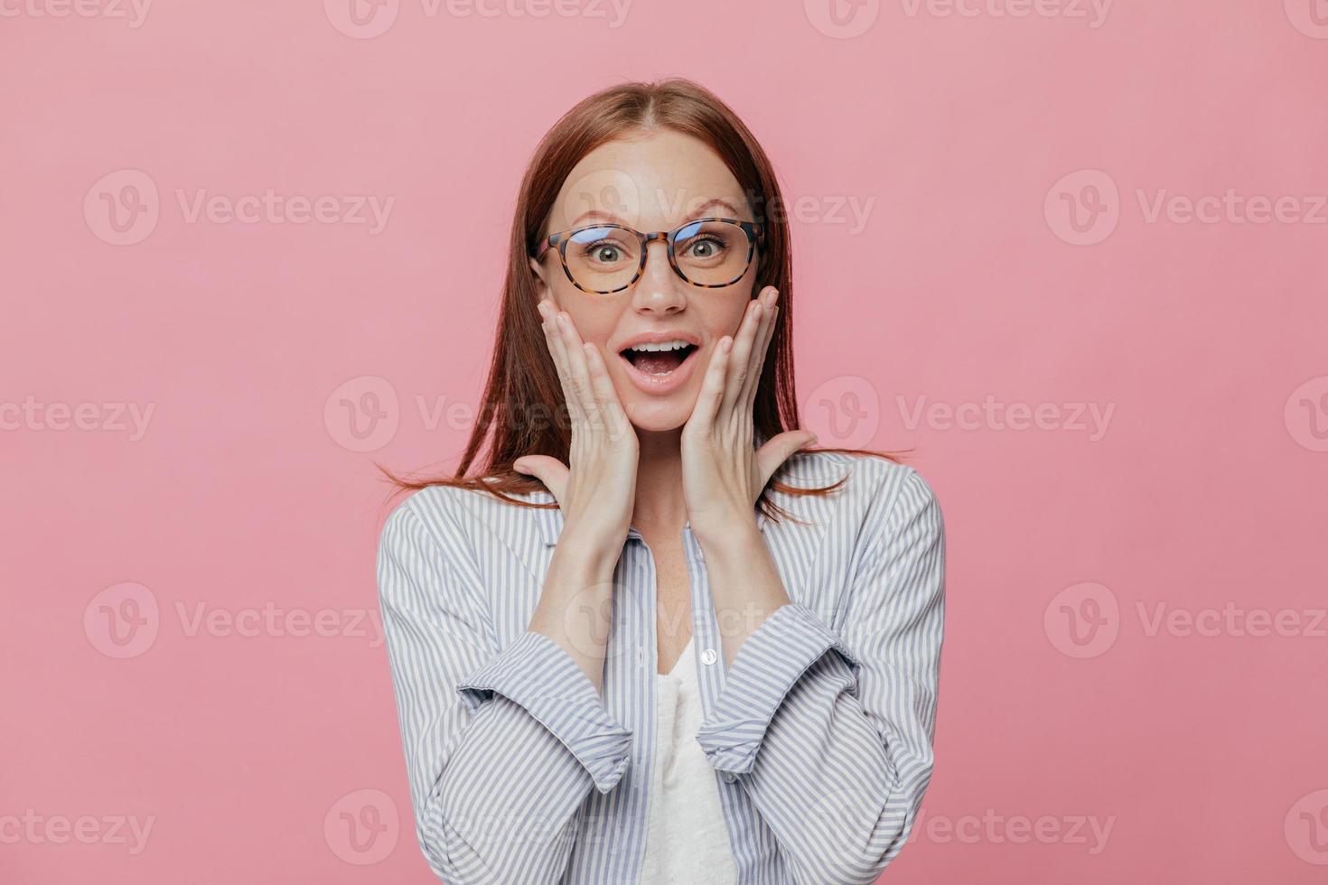 Omg, how nice Beautiful elegant female employee touches cheeks with both palms, wears spectacles, elegant shirt, cant believe in her promotion, recieves reward, isolated over pink background photo