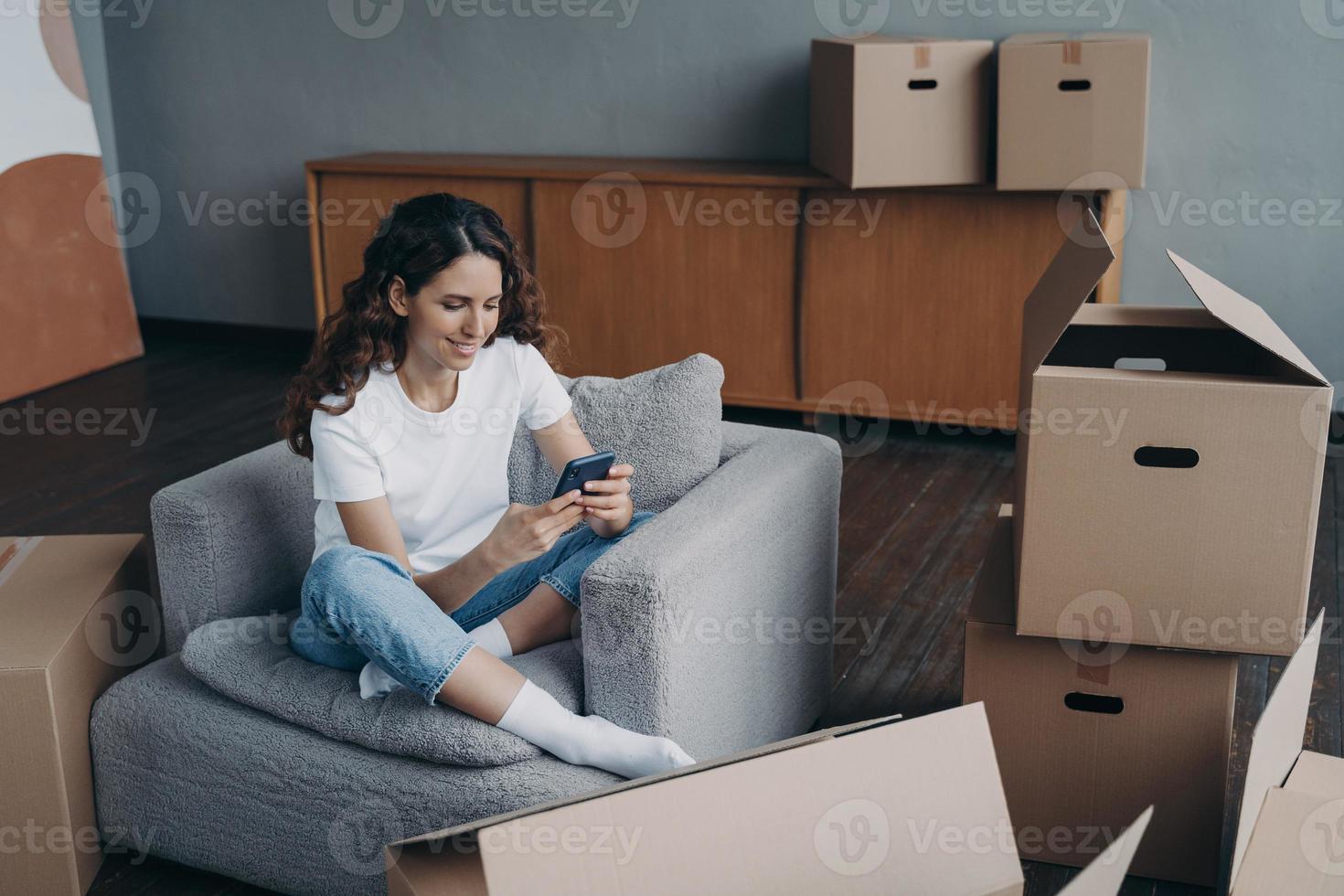 Smiling girl holding smartphone, selects moving company online for relocation, sitting with boxes photo