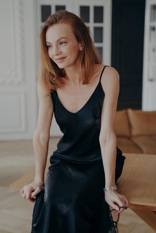 Attractive lady in black silk dress is sitting on table. Gorgeous woman in trendy cocktail dress. photo