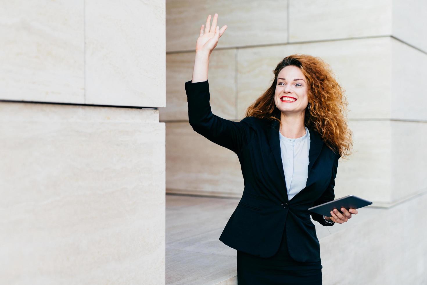 mujer de negocios sonriente y feliz vestida formalmente, sosteniendo en las manos una tableta moderna, saludando con la mano mientras notaba a su compañero, alegrándose de verlo. mujer con cabello rizado saludando foto