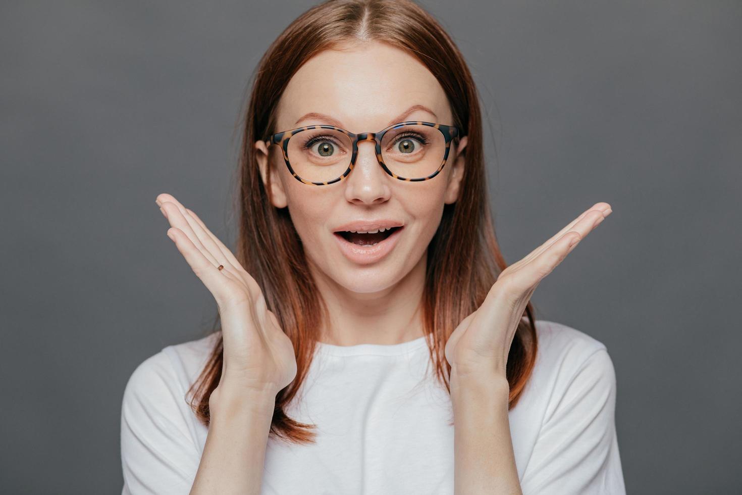 Wow, thats great Surprised pleased beautiful woman has pleasant appearance, spreads palms near face, opens mouth from wonder, dressed in casual t shirt, isolated over grey studio background photo