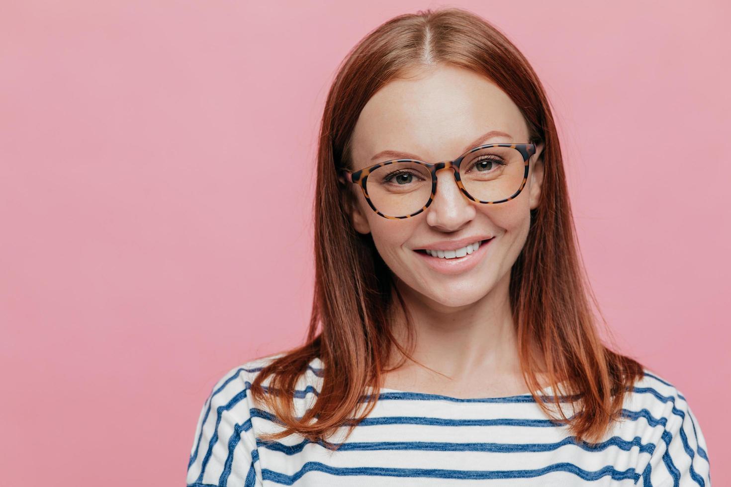 Close up shot of adorable young female student wears optical glasses, has tender smile, rejoices holidays wears striped clothes, models over pink background with copy space for your information. photo