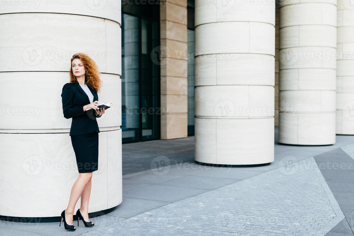 Beautiful woman with curly hair, slender legs, wearing black costume and high-heeled shoes, holding notebook in hands, arranging meeting with business partner standing near big office builduing photo