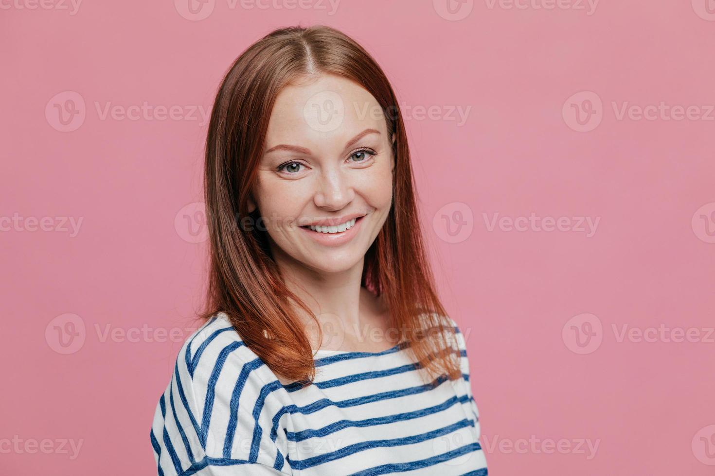 toma lateral de una mujer feliz de cabello castaño de aspecto agradable con cabello lacio, piel sana, sonrisa agradable, vestida con un suéter casual a rayas, aislada sobre fondo rosa. concepto de emociones foto