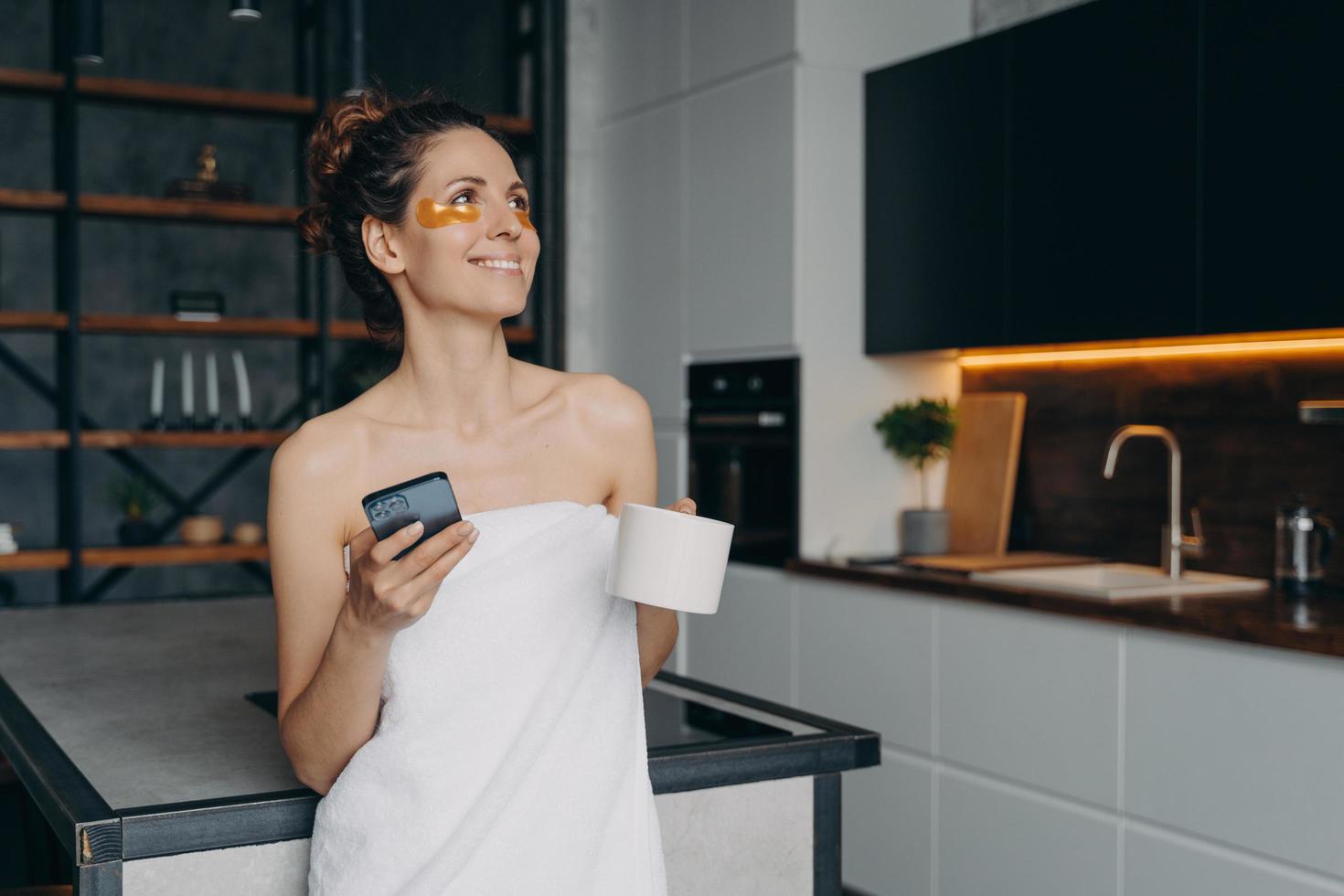 Happy woman with under eye patches holding smartphone and morning coffee got good news message photo