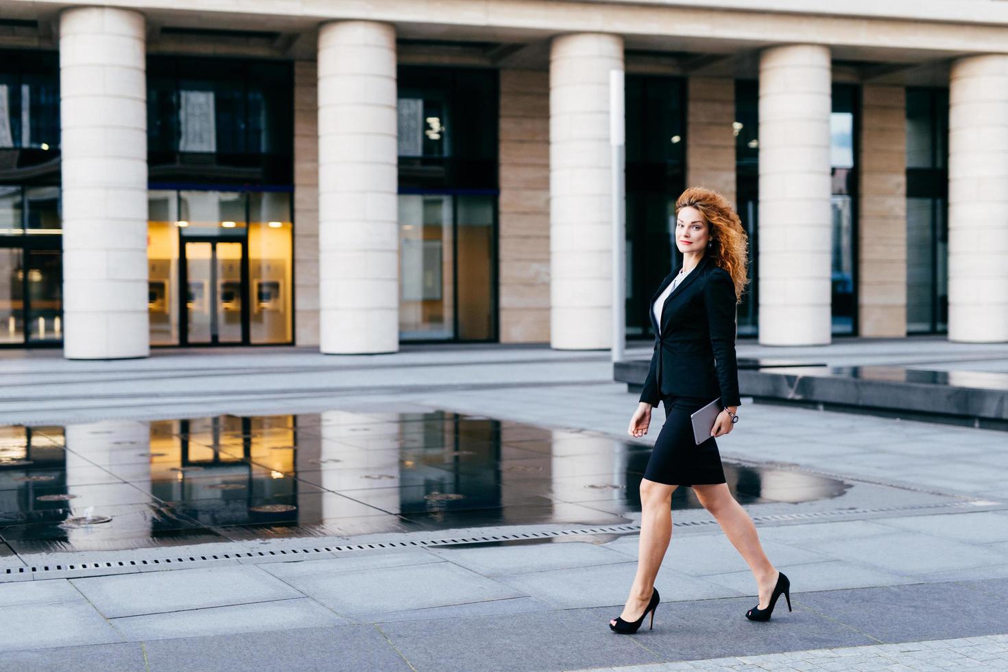 Elegant slim woman in black suit and high-heeled shoes, holding tablet in hands going for work. Businesswoman in motion. Confident beautiful young female in formal clothes near office builduing photo