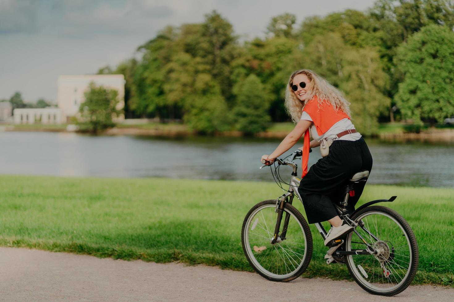 Photo of cheerful woman dressed casually, rides bicycle, looks aside, has happy expression, wears shades, poses near river, green lawn and trees, some buildings in background. People and rest