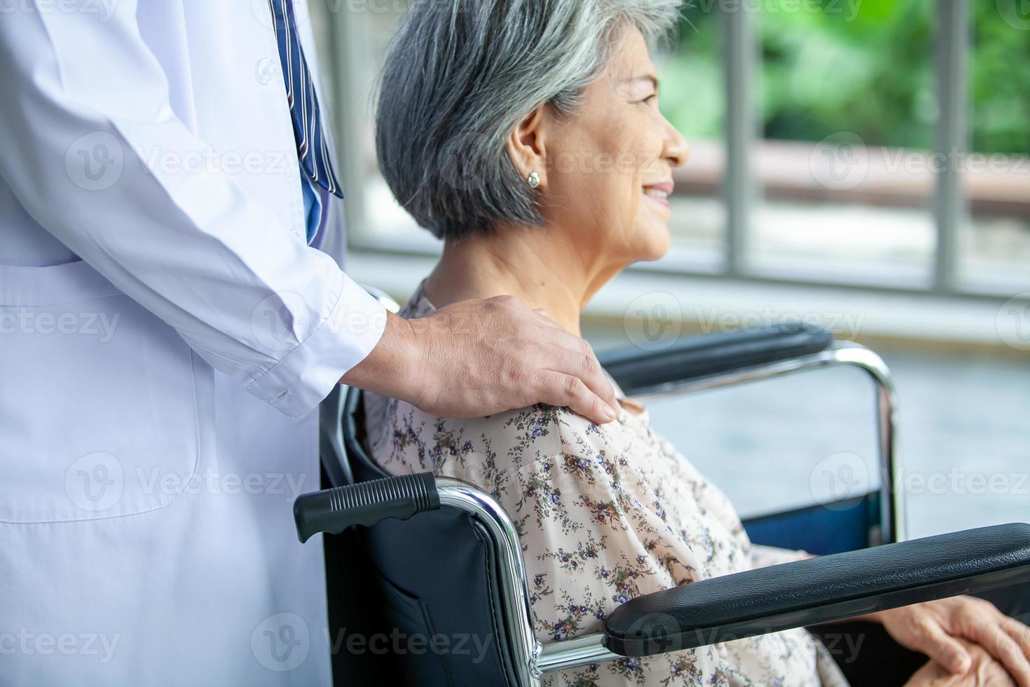 Asian doctor supporting and cheering up senior patient in wheelchair talking, smiling in comfort at home. Healthcare and medicine concept. photo