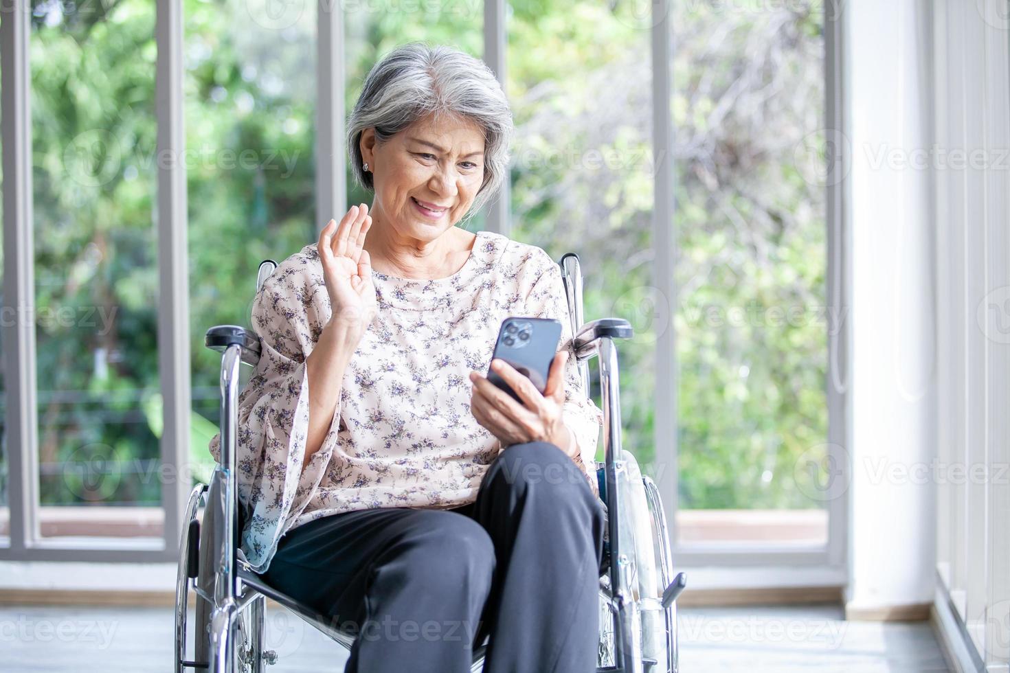 Happy elderly asian woman sitting on wheelchair and using smartphone for video call at home. photo