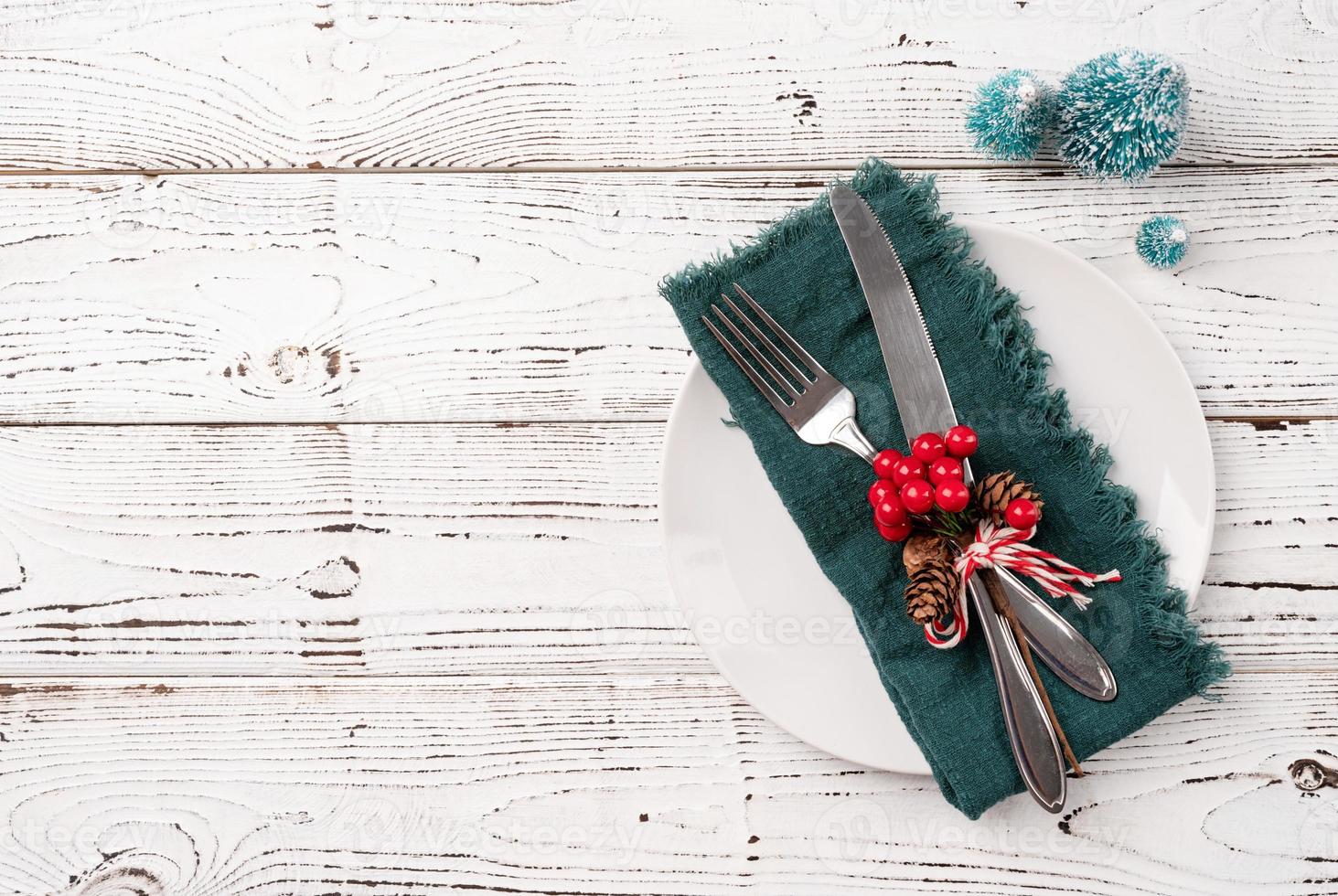 Christmas table setting with white dishware, silverware and red and green decorations on white wooden background. Top view. photo