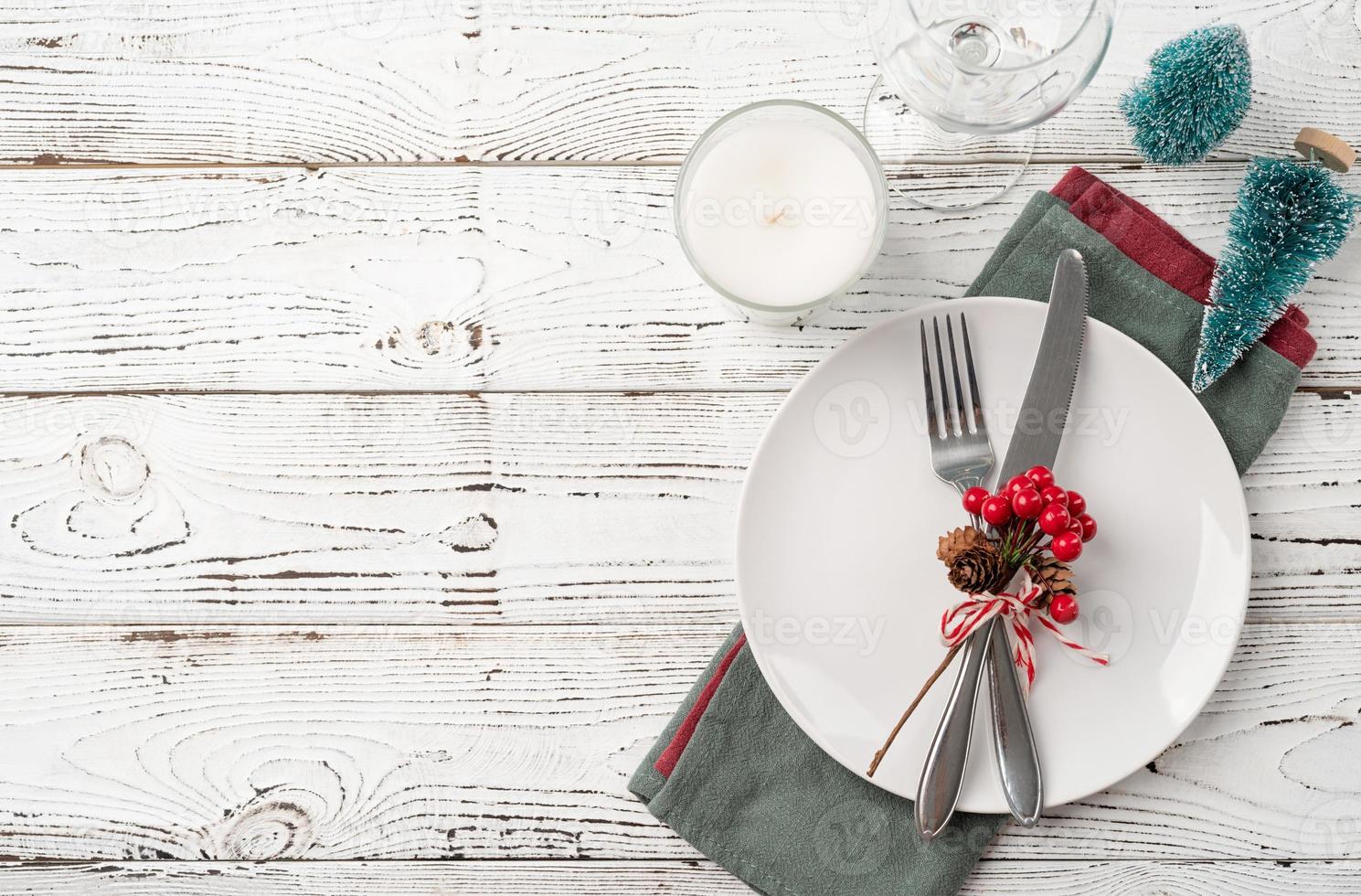 Christmas table setting with white dishware, silverware and red and green decorations on white wooden background. Top view. photo