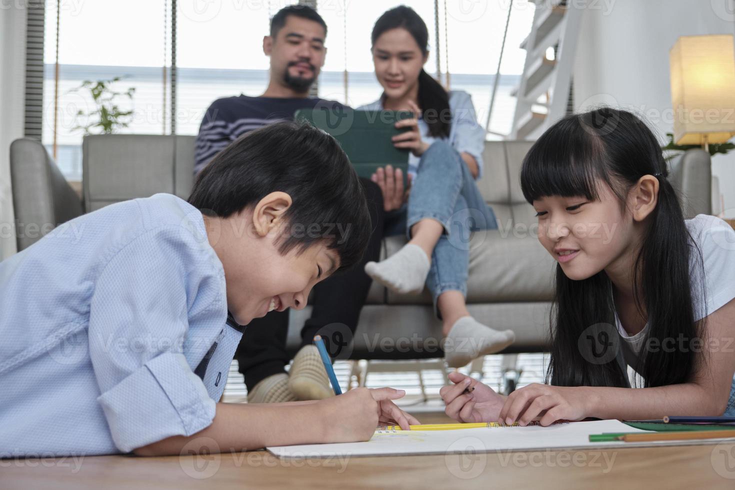 Asian Thai siblings are lying on living room floor, drawing homework with colored pencils together, parents leisurely relax on a sofa, lovely happy weekend activity, and domestic wellbeing lifestyle. photo