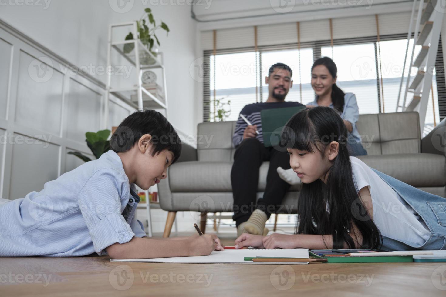 Asian Thai siblings are lying on living room floor, drawing homework with colored pencils together, parents leisurely relax on a sofa, lovely happy weekend activity, and domestic wellbeing lifestyle. photo