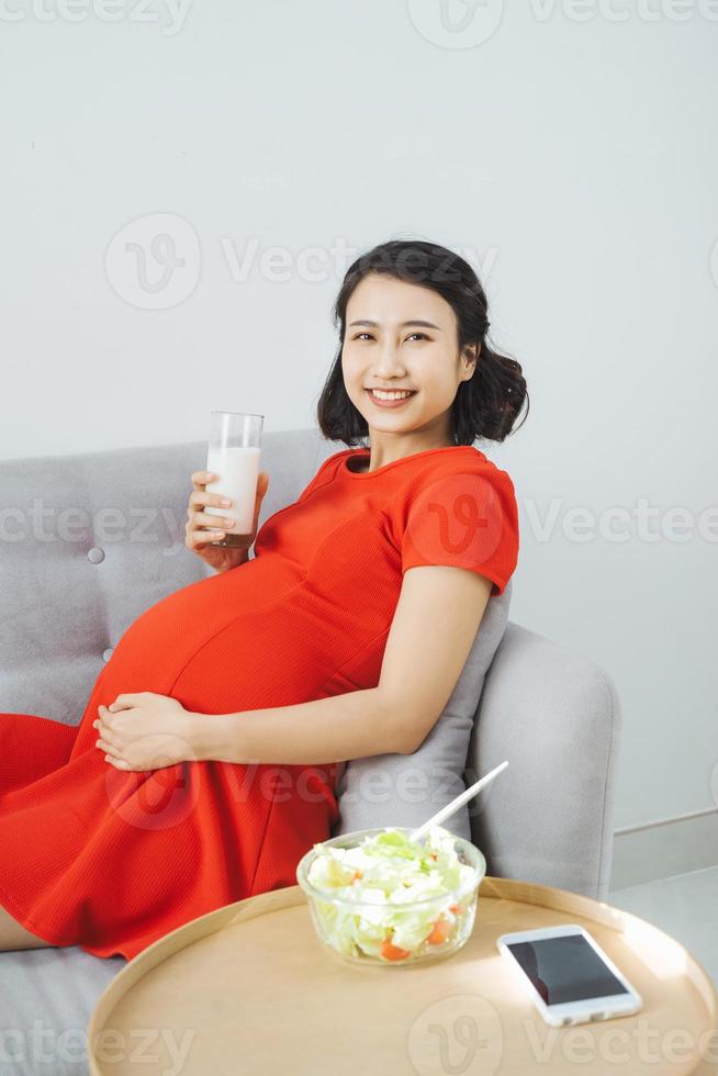 mujer embarazada asiática bastante joven comiendo ensalada y bebiendo leche cuando se sienta en el sofá. foto